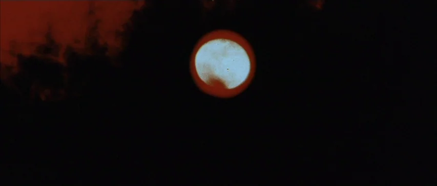 Messiah of Evil (1973) movie still: establishing — A deep black void fills the frame with a singular glowing celestial body. Dark red cloud wisps…