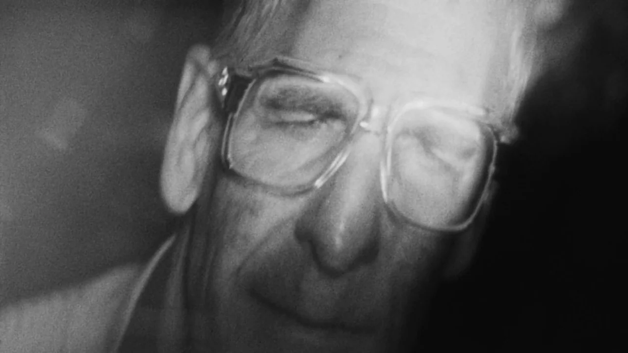 Divinity (2023) movie still: closeup — A dark indoor space contains an elderly man with his eyes closed. He wears thick-rimmed glasses while…