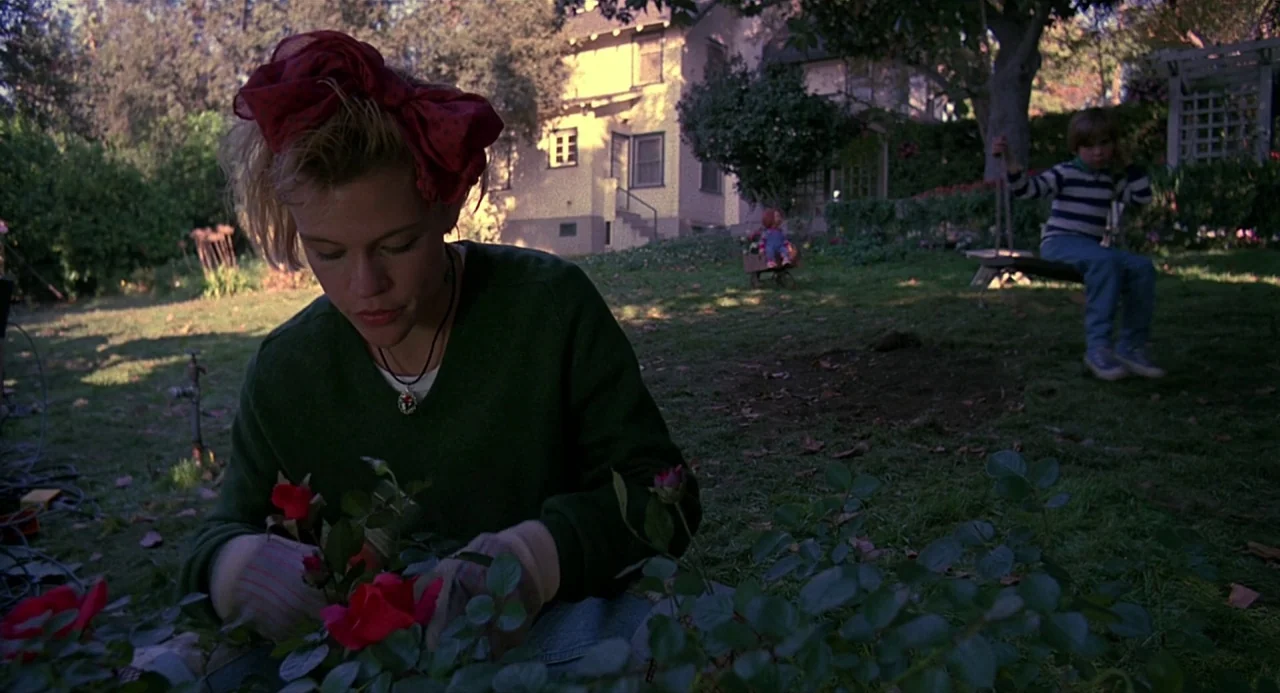 Child’s Play 2 (1990) movie still: medium — A sunny yard features a woman tending to red rose bushes in the foreground. A young boy sits on a swing…