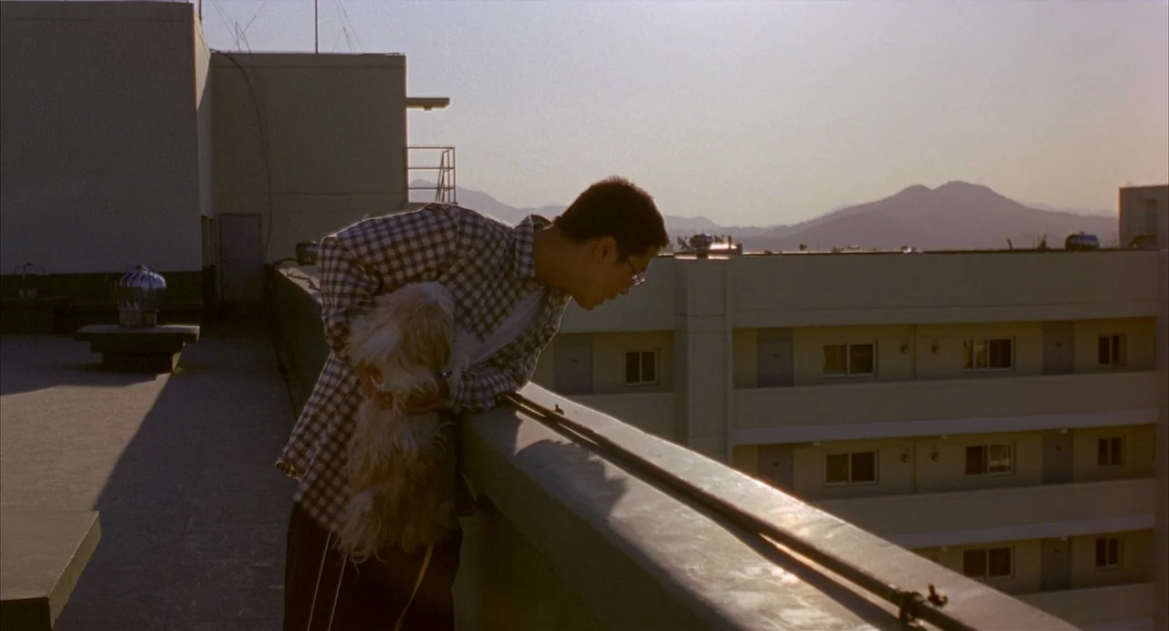 Barking Dogs Never Bite (2000) movie still: medium — A flat concrete building terrace features a low ledge and views of distant hills. A man holds a…