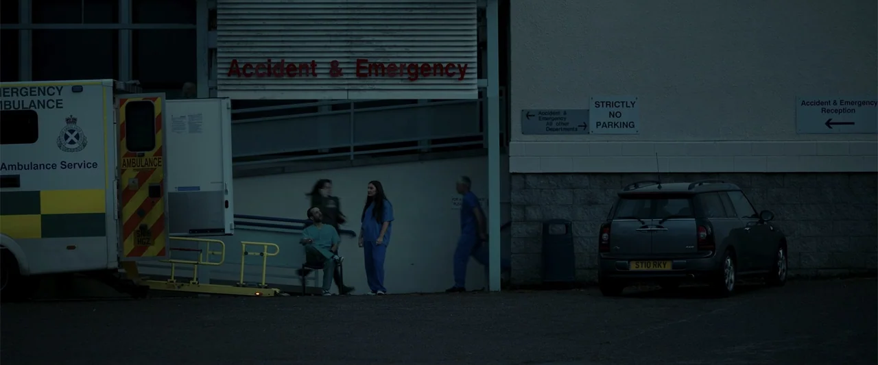 My Son (2021) movie still: wide — An exterior hospital emergency entrance at dusk features an ambulance parked beside the building. Medical staff in…