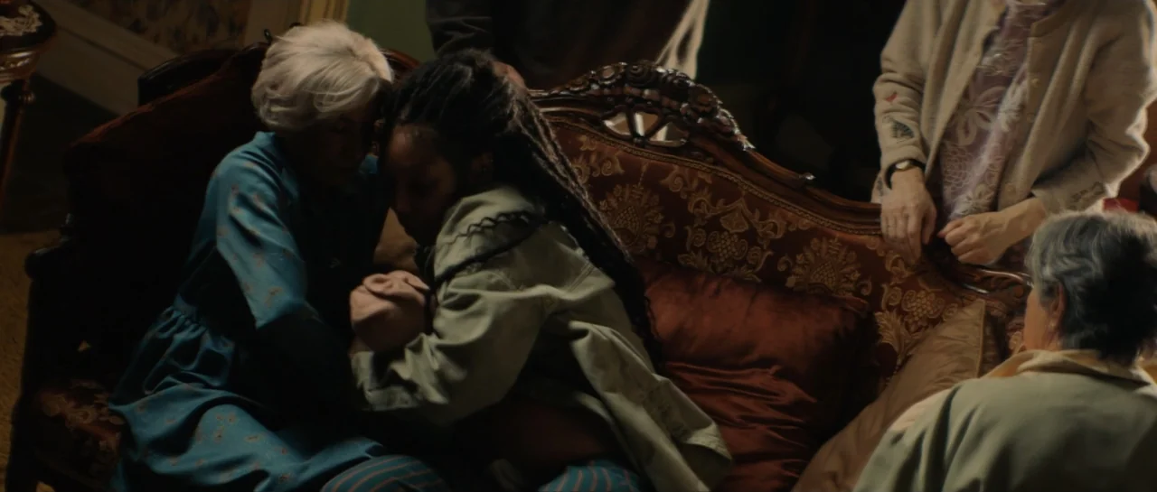 The Front Room (2024) movie still: medium — A dimly lit room features a dark wood ornate sofa with patterned fabric upholstery. A woman with long…