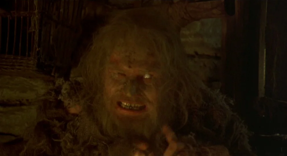 Monty Python and the Holy Grail (1975) movie still: closeup — A dark, cramped interior with rough wooden walls and shadows. A disheveled older man…