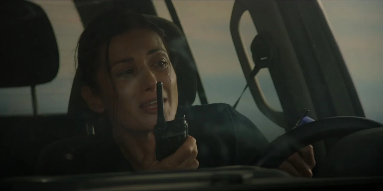 The Hunted (2024) movie still: medium — A woman sits in the driver's seat of a car during sunset. She speaks into a portable radio while holding the…