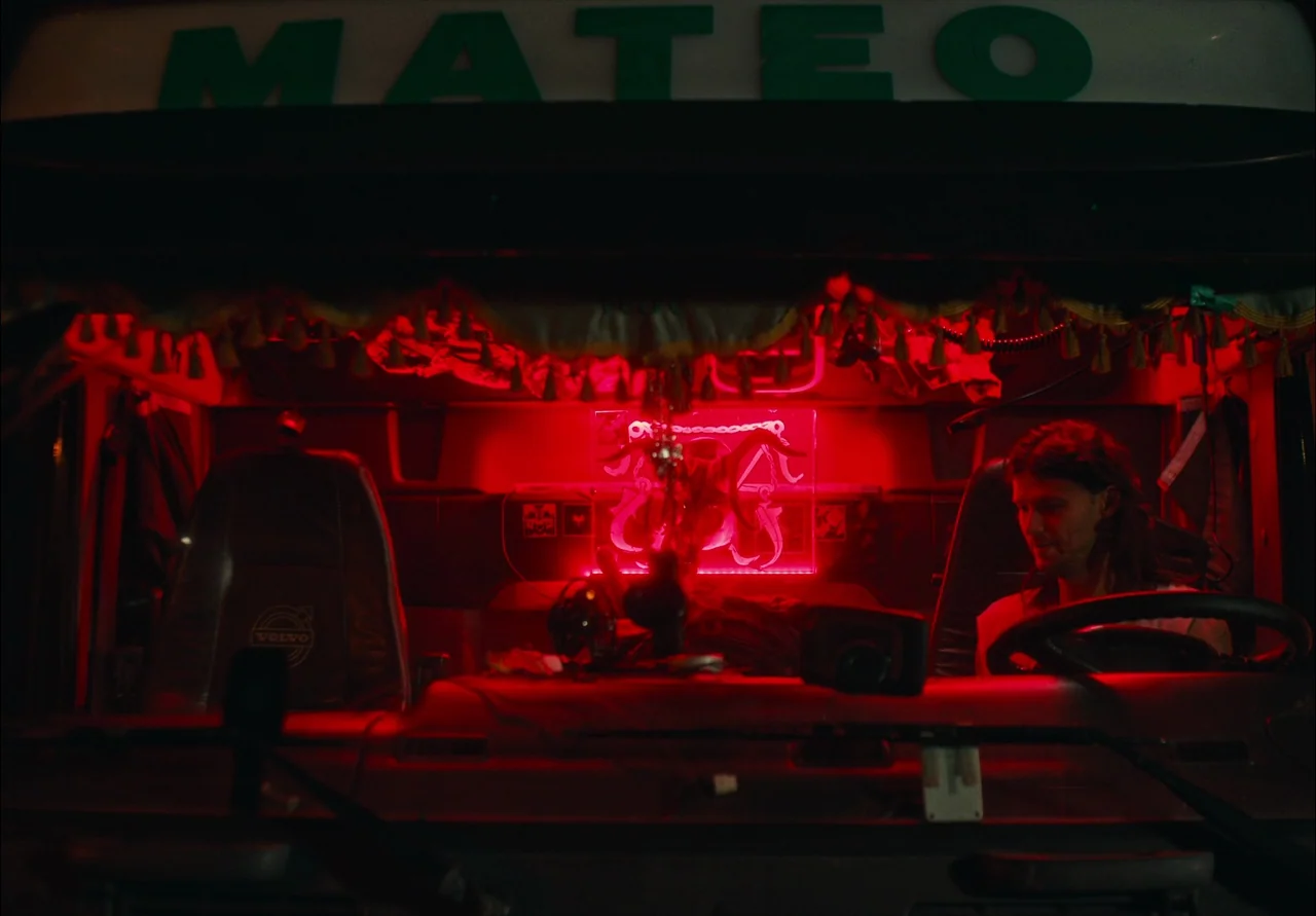 EO (2022) movie still: medium — The interior of a Volvo truck cab displays a bright red neon emblem. A man with shoulder-length hair sits behind the…