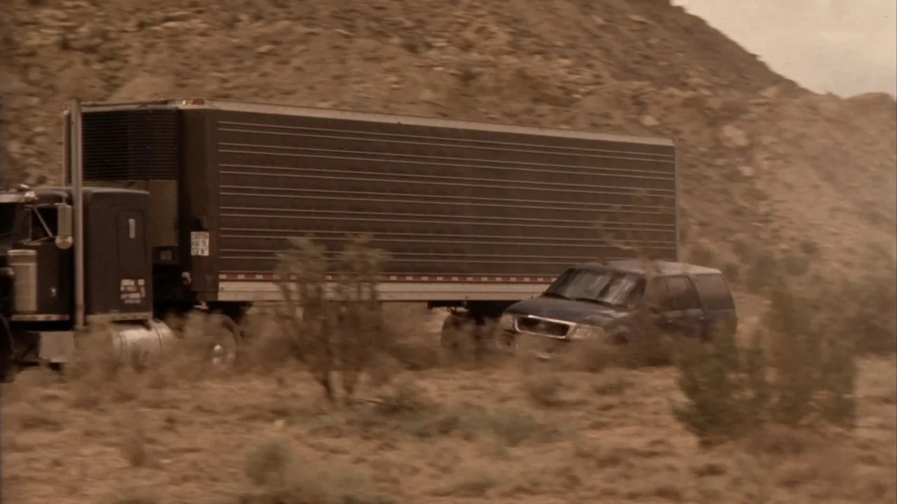Suspect Zero (2004) movie still: wide — A dark semi-truck and a dark SUV traverse a dusty, arid desert terrain. The vehicles move horizontally…
