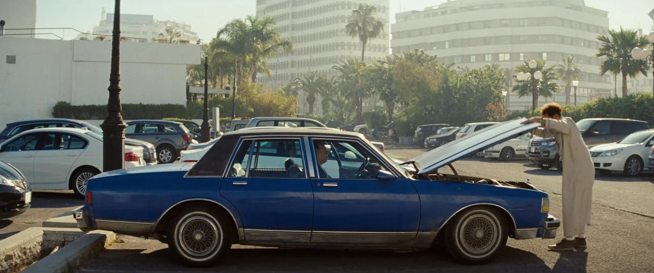 A Hologram for the King movie still: wide — A sunny outdoor parking lot is filled with numerous cars and distant city buildings. A man in a long…