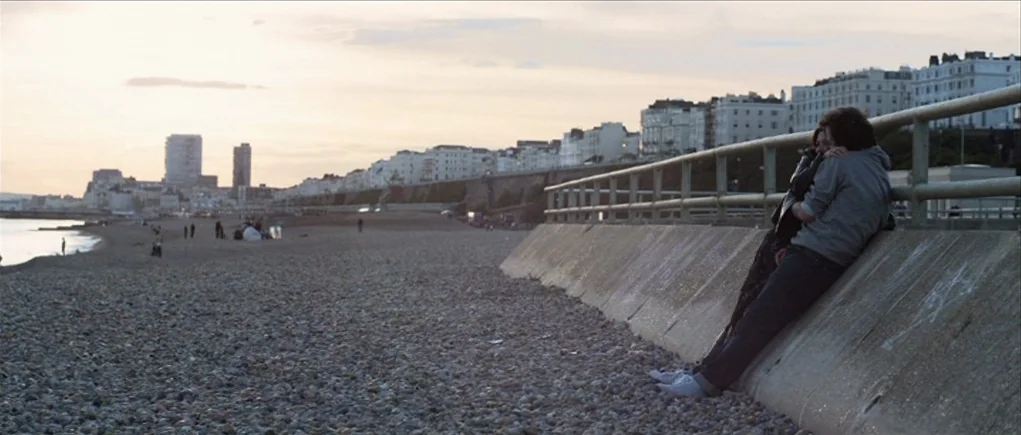 Down Terrace (2009) movie still: wide — A wide pebble beach stretches along a coastline with a row of white buildings. Two people lean against a low…