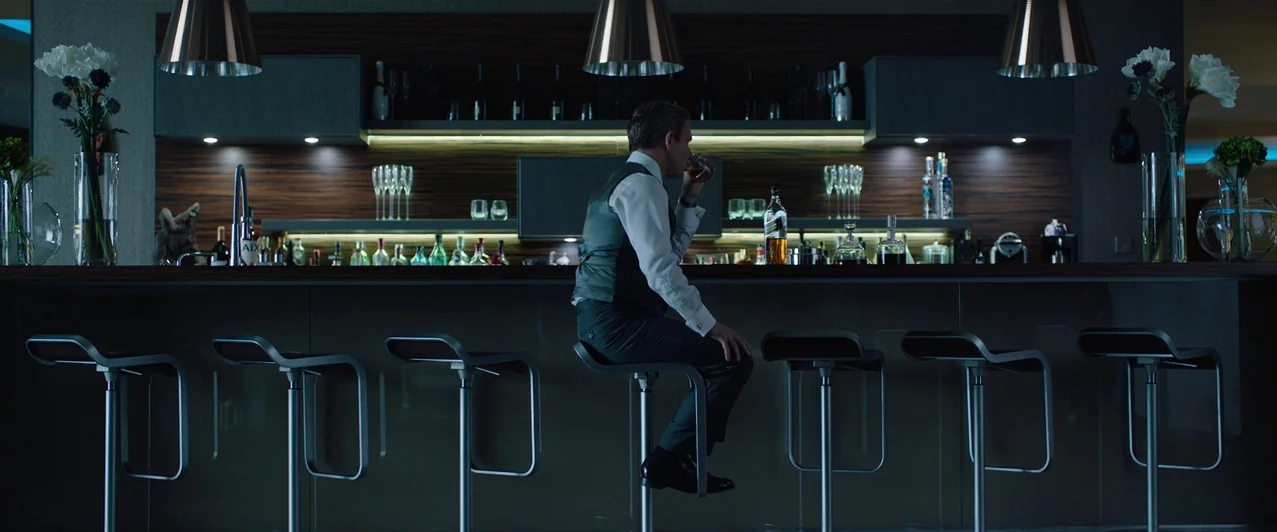 Ghost Stories (2017) movie still: wide — A contemporary bar features dark wood shelving, modern stools, and minimalist pendant lights. A man sits…