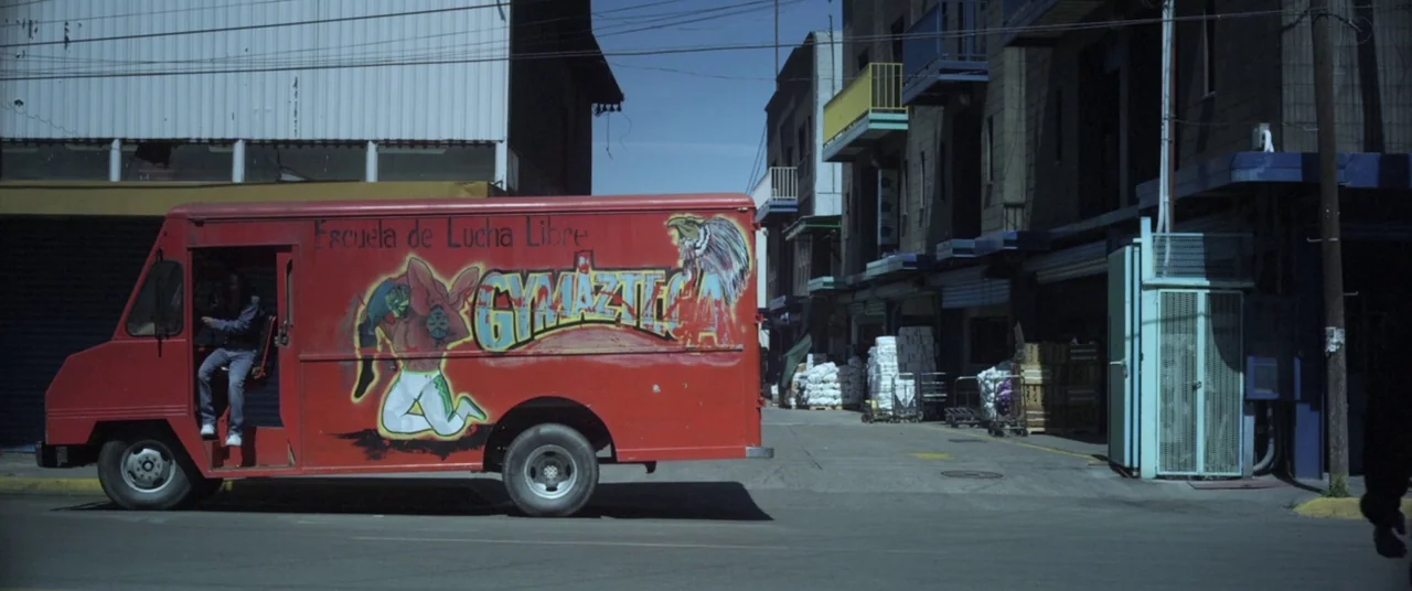 Here Comes The Devil (2012) movie still: wide — A vibrant red van with luchador wrestling murals parked on a concrete city street. A driver sits…