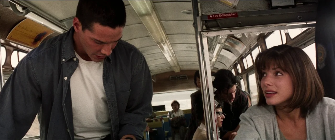 Speed (1994) movie still: medium — The interior of a bus has aged vinyl seats and a metal partition. A man in a denim shirt and a woman in a grey…