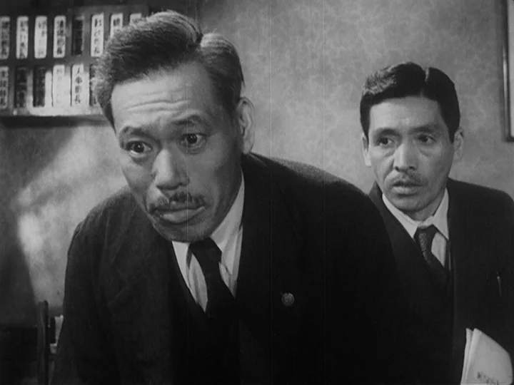 Ikiru (1952) movie still: medium — An interior office space features a wall board with text and textured wallpaper. Two men in suits look toward an…