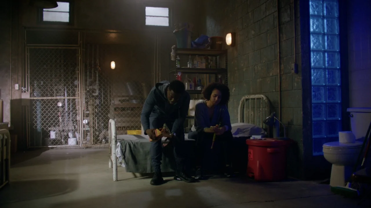 Die Hart (2023) movie still: fullbody — A concrete cell contains two cots, a red biohazard bin, and a tiled wall. A man and a woman sit on their…