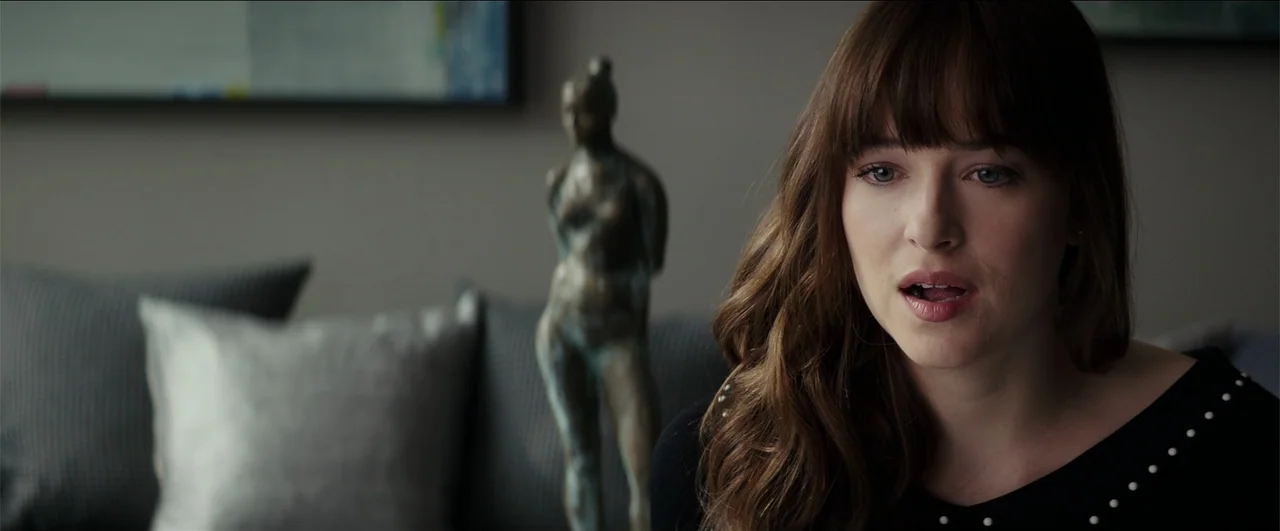 Fifty Shades Freed (2018) movie still: closeup — A living room interior features a blurred bronze sculpture in the background. A woman in a black…