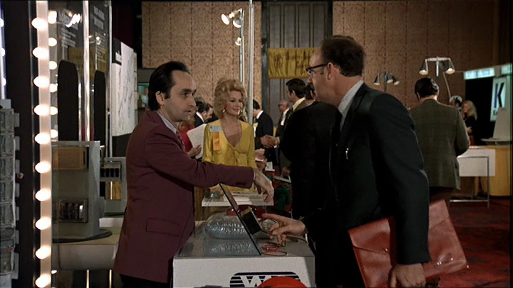 The Conversation (1974) movie still: medium — An indoor trade show floor features patterned wallpaper and multiple exhibition booths with lighting…
