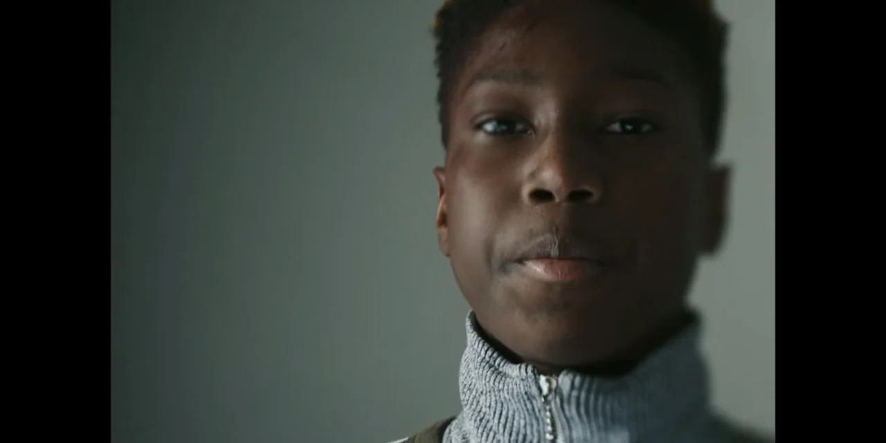 Autumn Beat (2022) movie still: closeup — A close-up of a young person in a room with a plain grey wall background. The subject faces the camera…
