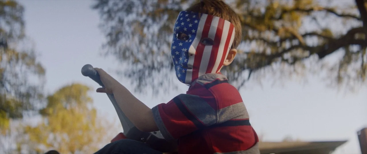 Assassination Nation (2018) movie still: medium — An outdoor neighborhood setting features a child amidst blurred trees during golden hour. The…