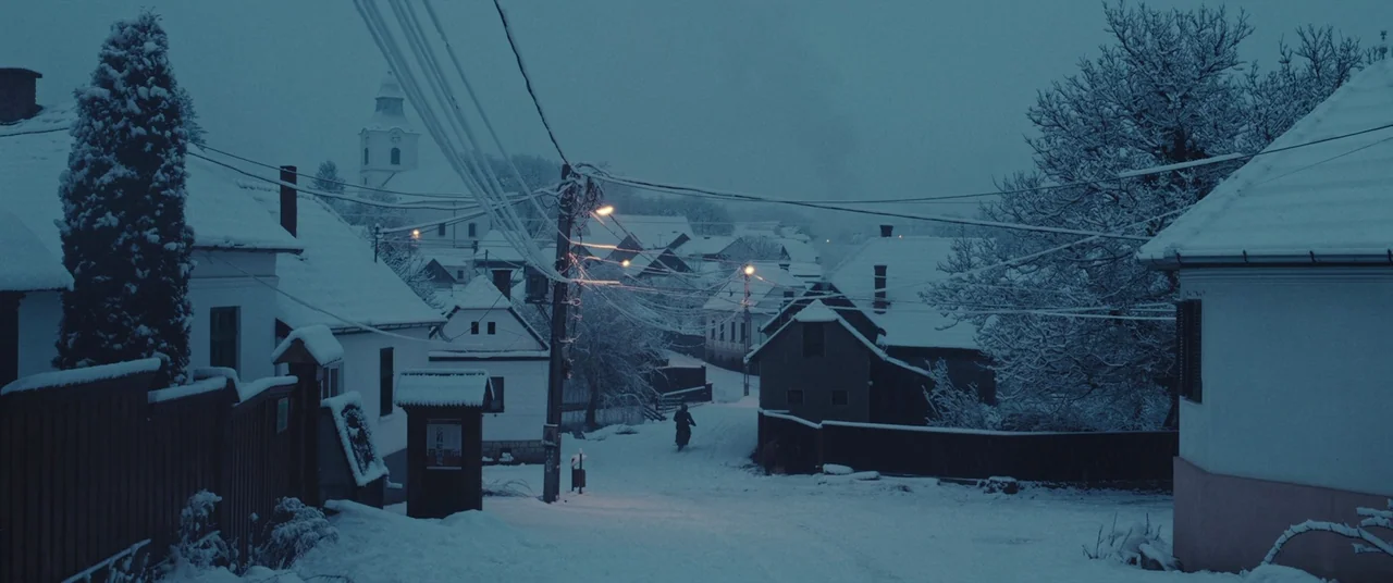 R.M.N. (2022) movie still: wide — A snow-covered village street at dusk with white cottages and bare trees. A lone bicyclist travels along the…