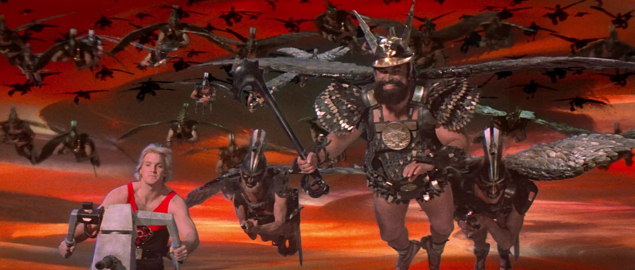 Flash Gordon (1980) movie still: wide — A group of winged warriors flies through a bright red, abstract gaseous sky. A blond man maneuvers a small…