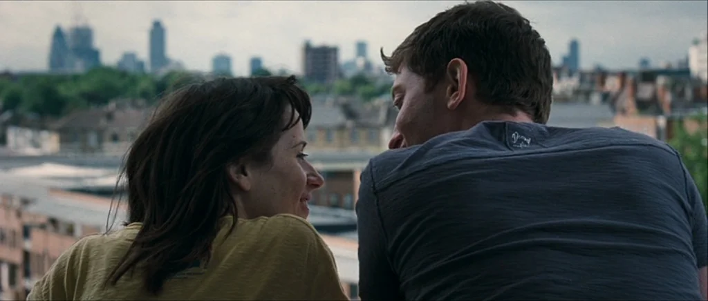Happy-Go-Lucky (2008) movie still: medium — A man and woman lean against a rooftop ledge overlooking a dense city skyline. They face each other in…