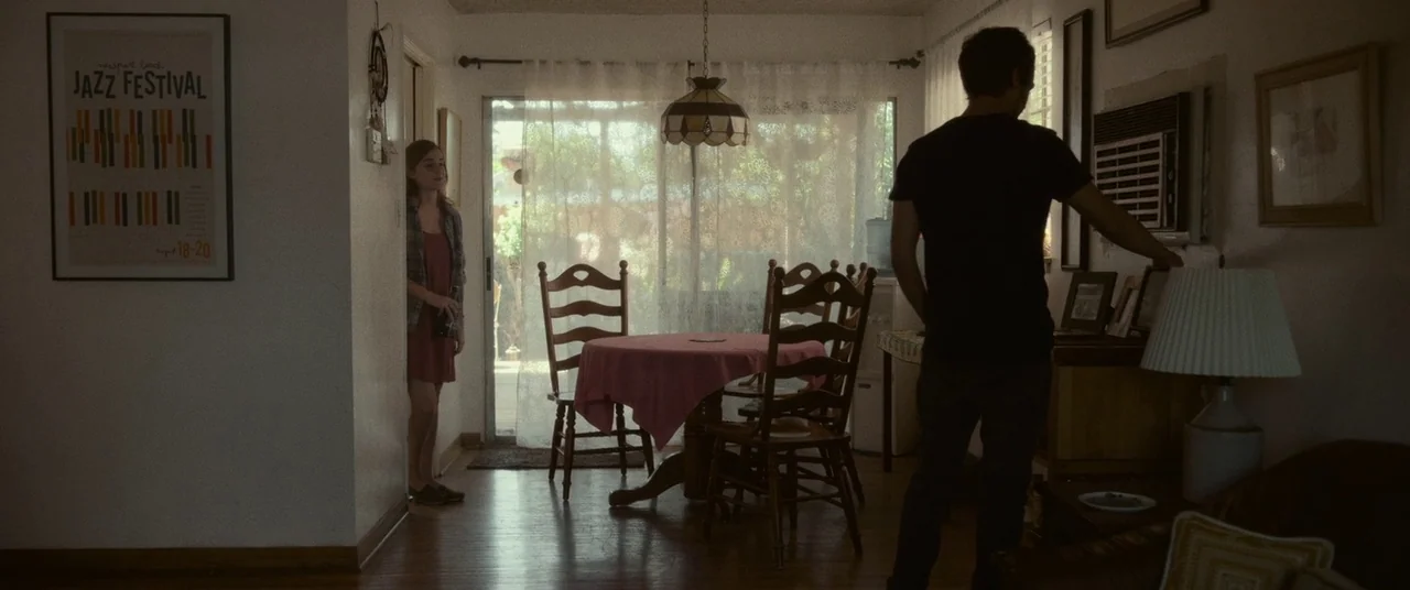 The Circle (2017) movie still: wide — A residential room features a small table with a pink cloth and wooden chairs. A man stands by the wall unit…