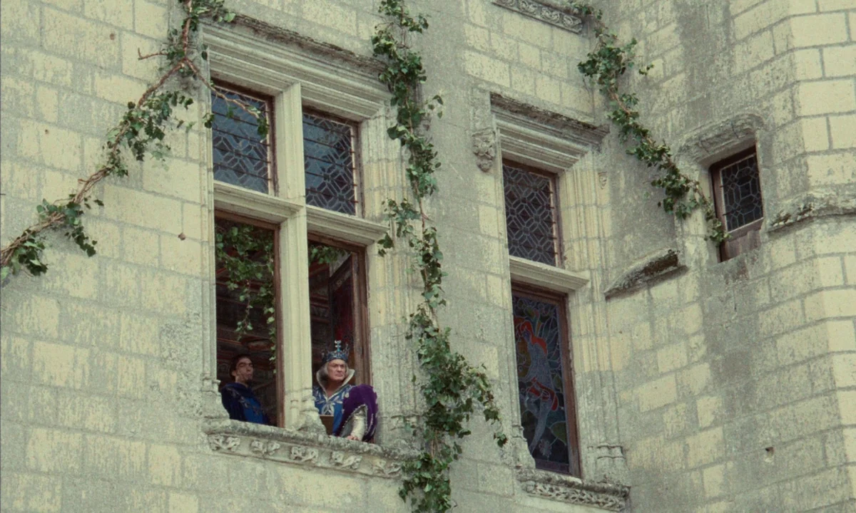 Donkey Skin (1970) movie still: wide — A stone castle exterior features carved window frames and climbing ivy vines. An elderly king in a crown and…
