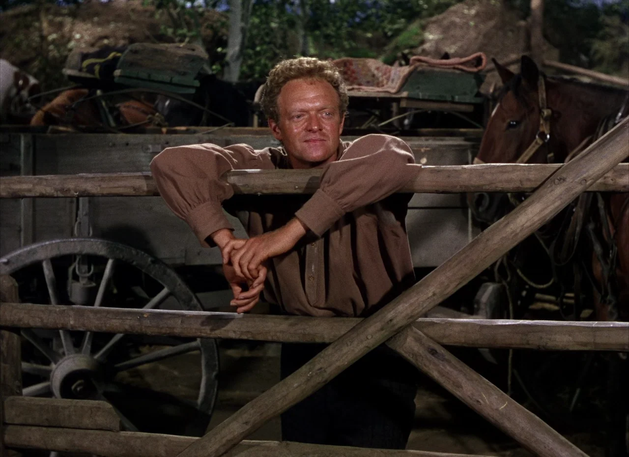 Shane (1953) movie still: medium — A rural setting features a wooden fence placed in front of a heavy wagon. A man rests his arms on the rail while…