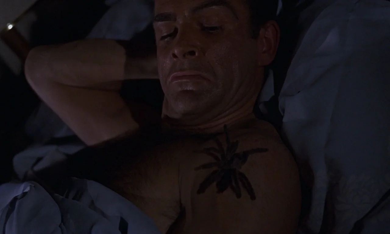 Dr. No (1962) movie still: closeup — A man lies on a bed with light blue sheets in a dim room. A large tarantula sits motionless on the man's bare…