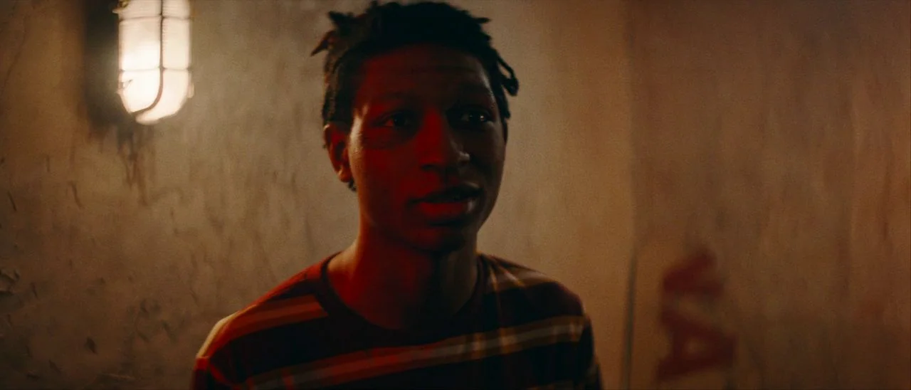 Archenemy (2020) movie still: medium — A dimly lit industrial hallway features textured concrete walls and a caged wall light. A young man wearing a…
