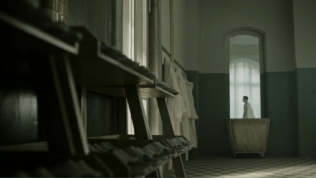 A Cure for Wellness (2017) movie still: wide — A utilitarian laundry room with tiled floors and shelves of folded linens in foreground. A distant…