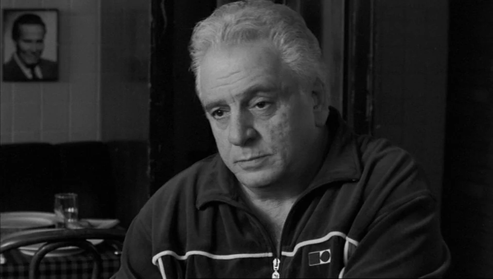 Coffee and Cigarettes (2003) movie still: closeup — An older man sits at a table in a dimly lit restaurant. A small framed portrait hangs on the…