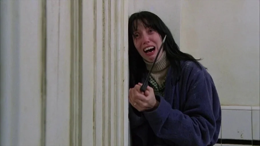 The Shining (1980) movie still: medium — A cramped tiled bathroom interior with plain white walls and vertical molding. A distraught woman clutches…