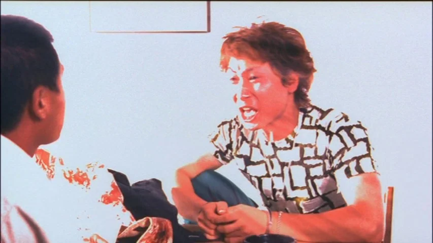 Save The Green Planet! (2003) movie still: medium — Two men sit at a simple wooden table against a plain white wall. One man leans forward with an…