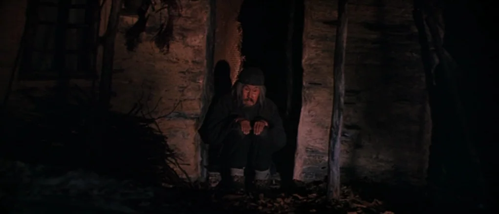 Dersu Uzala (1975) movie still: medium — A man in a dark tunic sits against a rough stone wall at night. The man remains still while resting his…