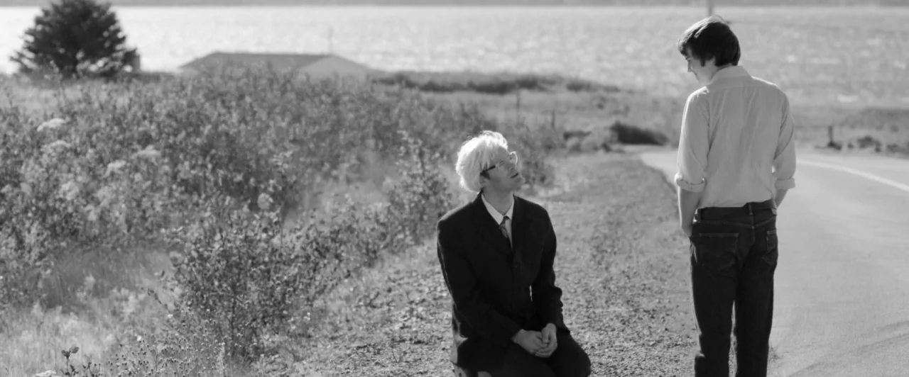 Weirdos (2016) movie still: fullbody — A rural road runs parallel to a body of water with tall dry grasses. A man kneels in the dirt as a second man…