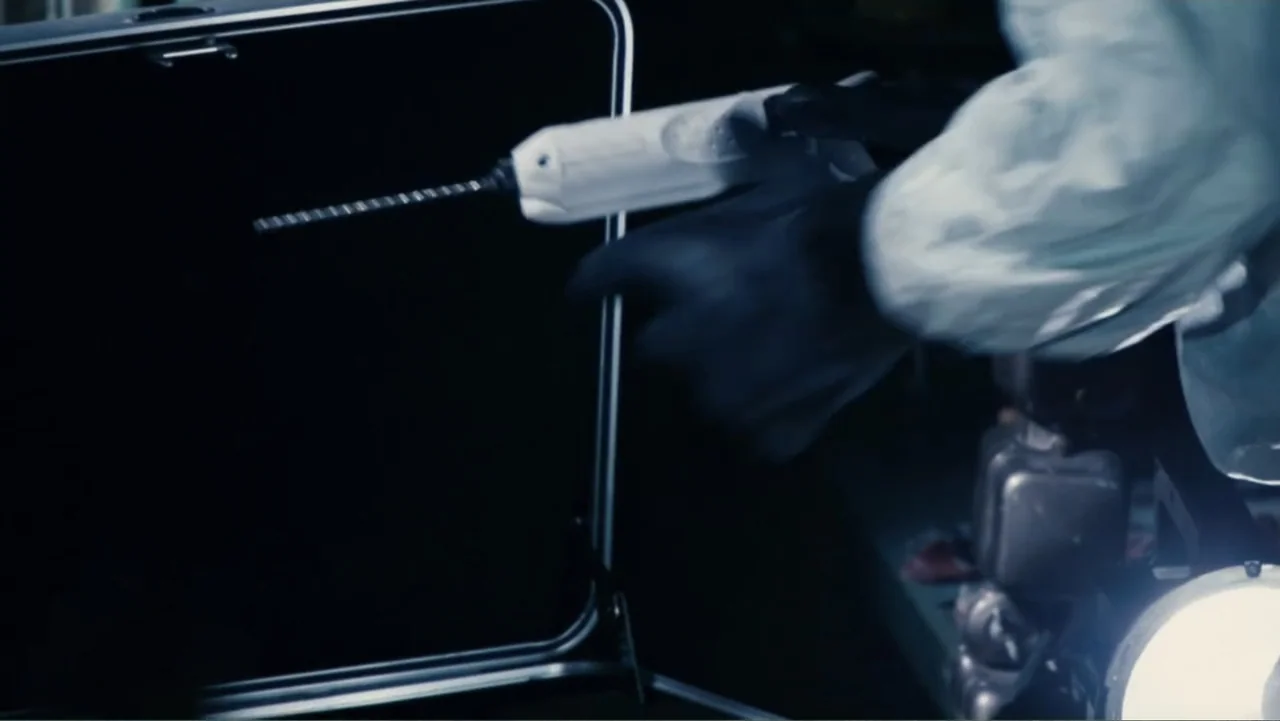 Quarantine (2008) movie still: closeup — A dimly lit industrial space contains a person in protective gear. The individual holds a white electric…