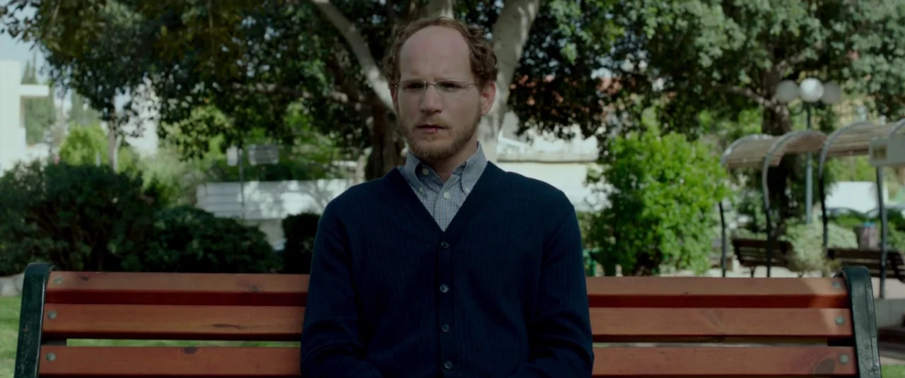 Big Bad Wolves (2013) movie still: medium — A man sits on a wooden slatted bench in an outdoor urban park area. Green trees and foliage provide a…