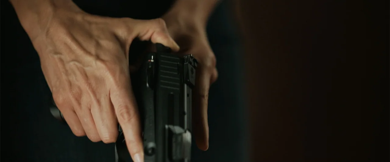 The 355 (2022) movie still: closeup — A close view shows two hands manipulating a modern black semi-automatic pistol. The subject is out of frame…