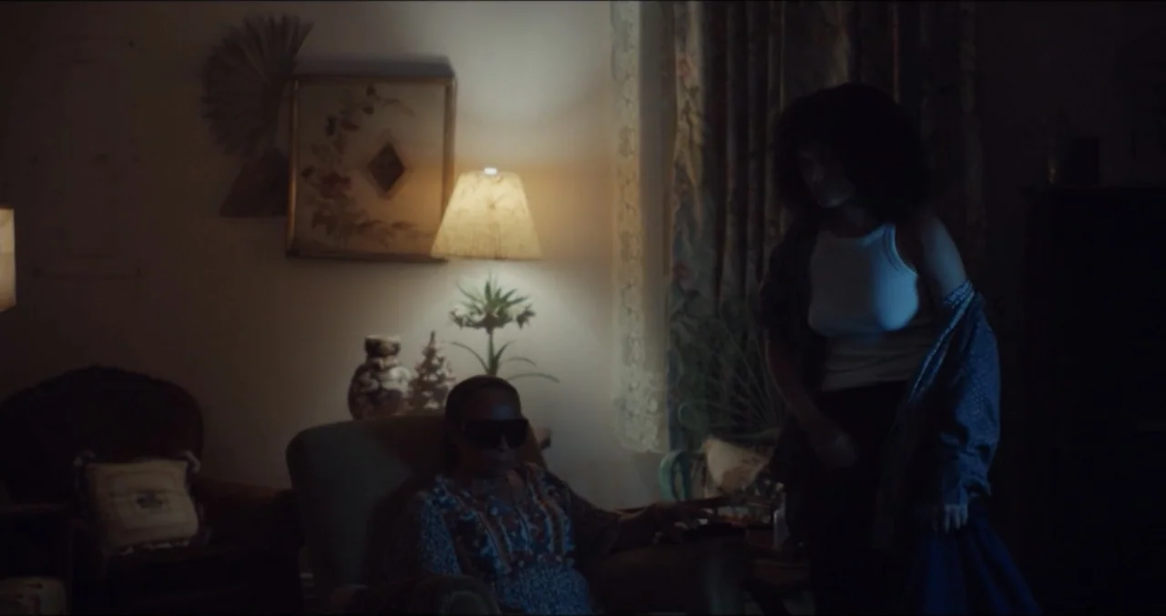 Eye for an Eye (2025) movie still: medium — A dimly lit living room features patterned wallpaper and a vintage table lamp. One woman sits in an…