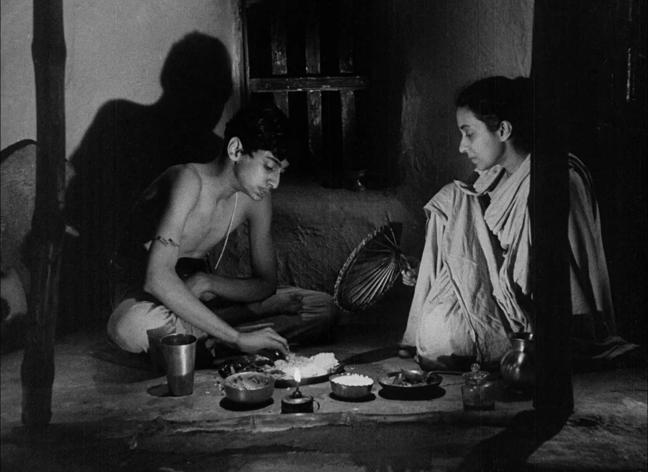 Aparajito (1956) movie still: medium — A dimly lit room with earthen walls and a single flickering candle on the floor. A shirtless young man eats…