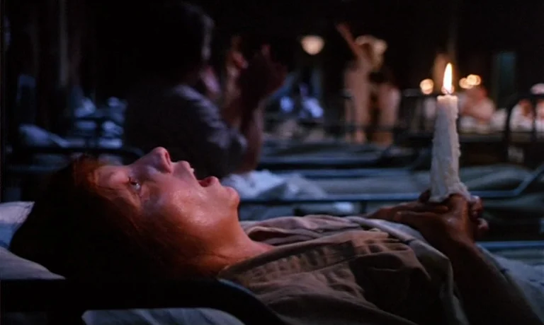 Katie Tippel (1975) movie still: medium — A dark hospital ward contains rows of metal bed frames occupied by patients. A woman lies in a bed with…