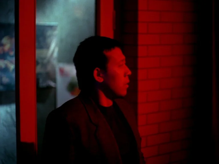 Black Snow (1990) movie still: medium — A man stands outdoors next to a red brick wall beside a glass door. A strong red light illuminates his…