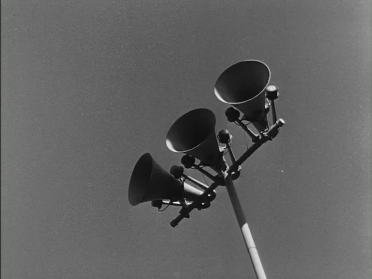 The Killing (1956) movie still: closeup — A cluster of three metal megaphone speakers mounted to a single vertical pole. The assembly is silhouetted…