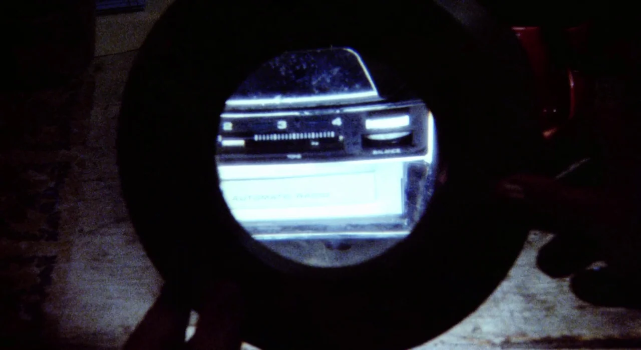 White Of The Eye (1987) movie still: closeup — A dark environment frames a vintage radio tuner seen through a handheld magnifying glass. The…