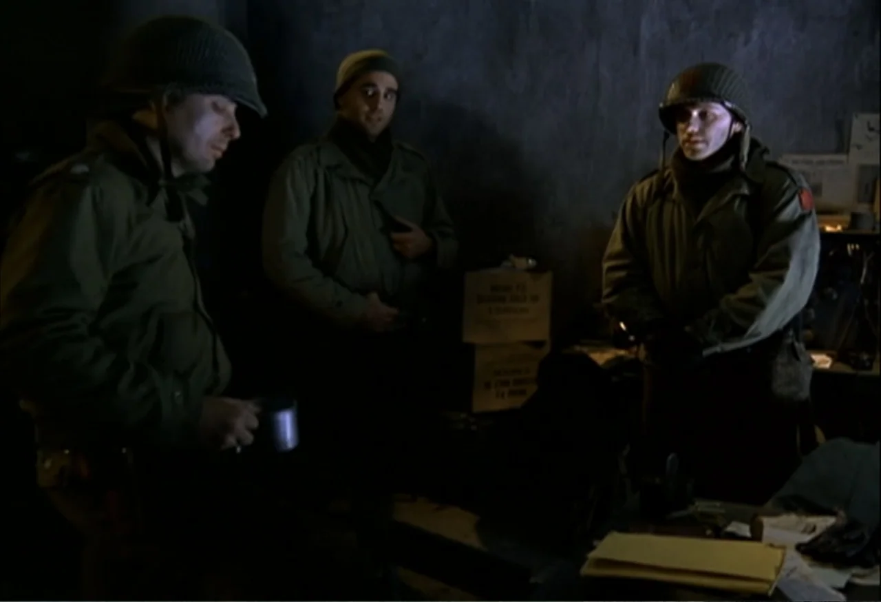When Trumpets Fade (1998) movie still: medium — A dark military bunker contains three soldiers standing near a cluttered desk. The men occupy the…