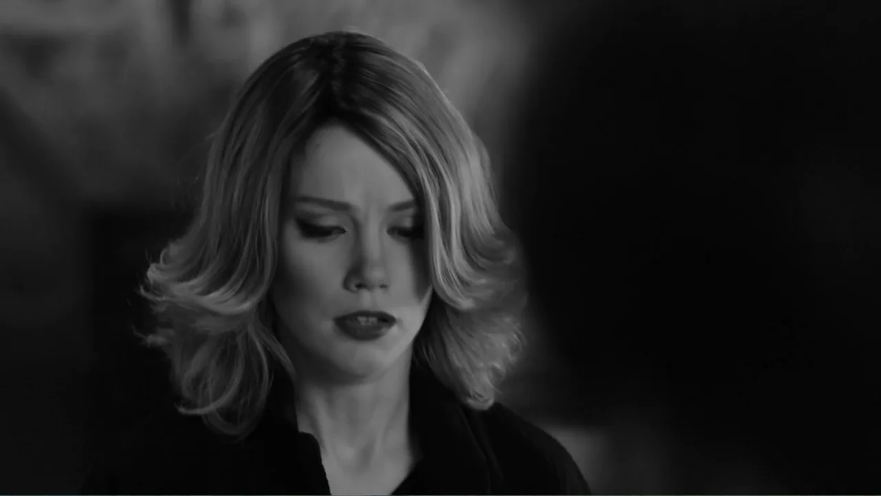 Creditors (2025) movie still: closeup — An interior space with indistinct dark surroundings. A woman with wavy blonde hair looks down with her eyes…