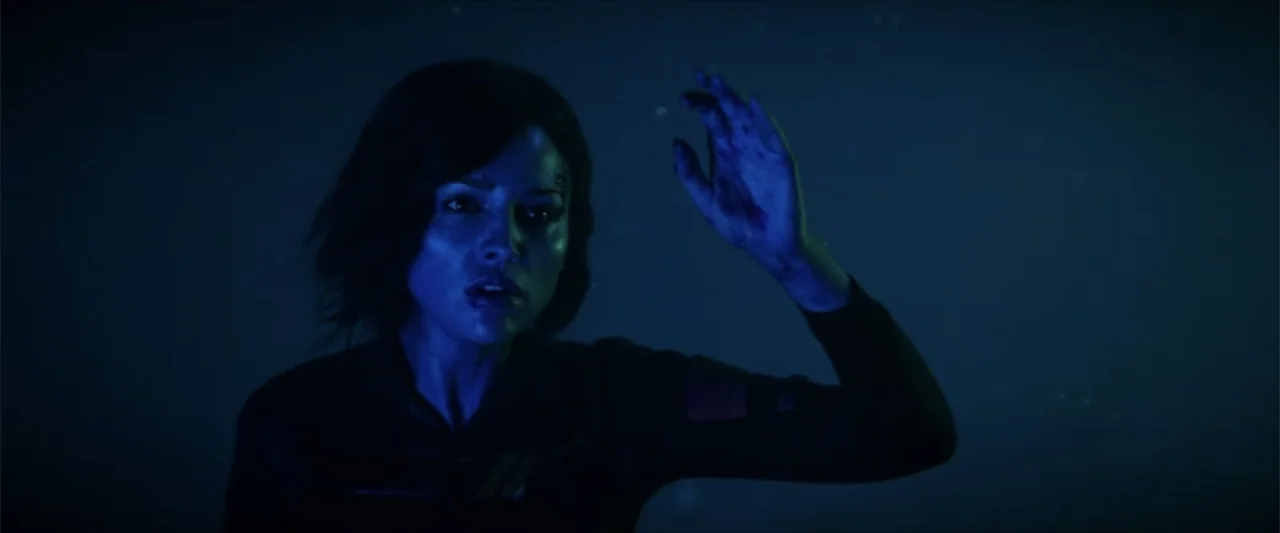 Ash (2025) movie still: medium — A dimly lit space features a woman surrounded by scattered glowing floating debris particles. She reaches out with…