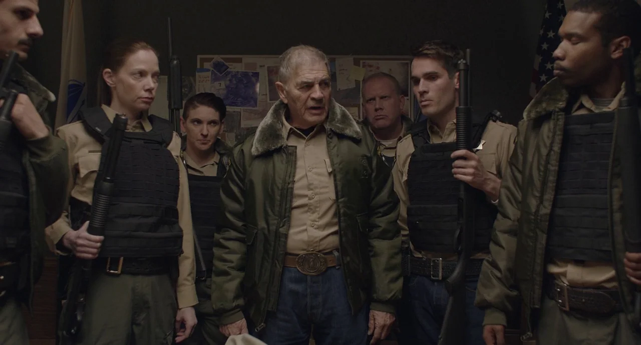 The Wolf of Snow Hollow (2020) movie still: medium — A group of police officers stands inside a dimly lit office with evidence pinned up. They hold…