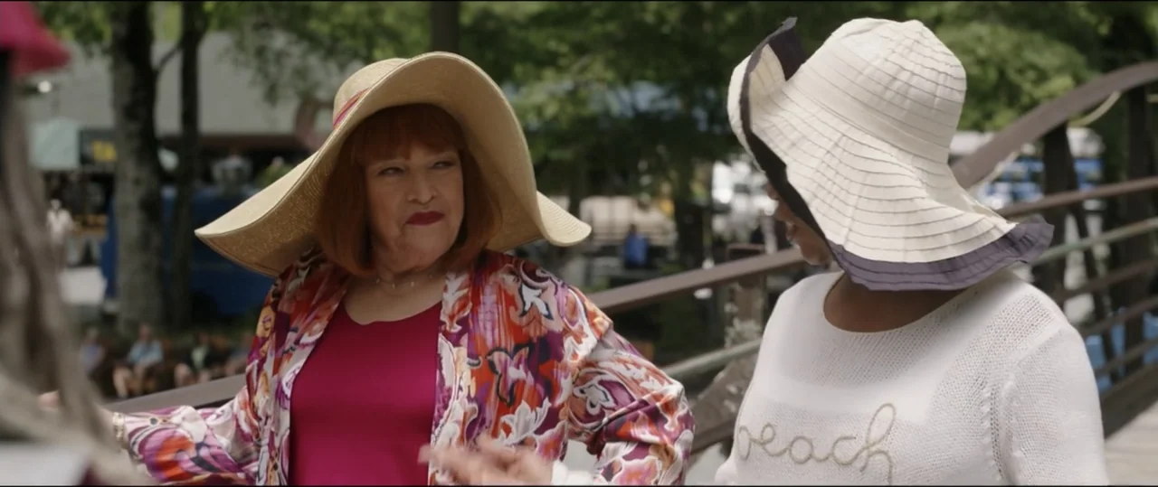 Summer Camp (2024) movie still: medium — A wooden pedestrian bridge in an outdoor park setting during the day. Two women in summer hats stand close…