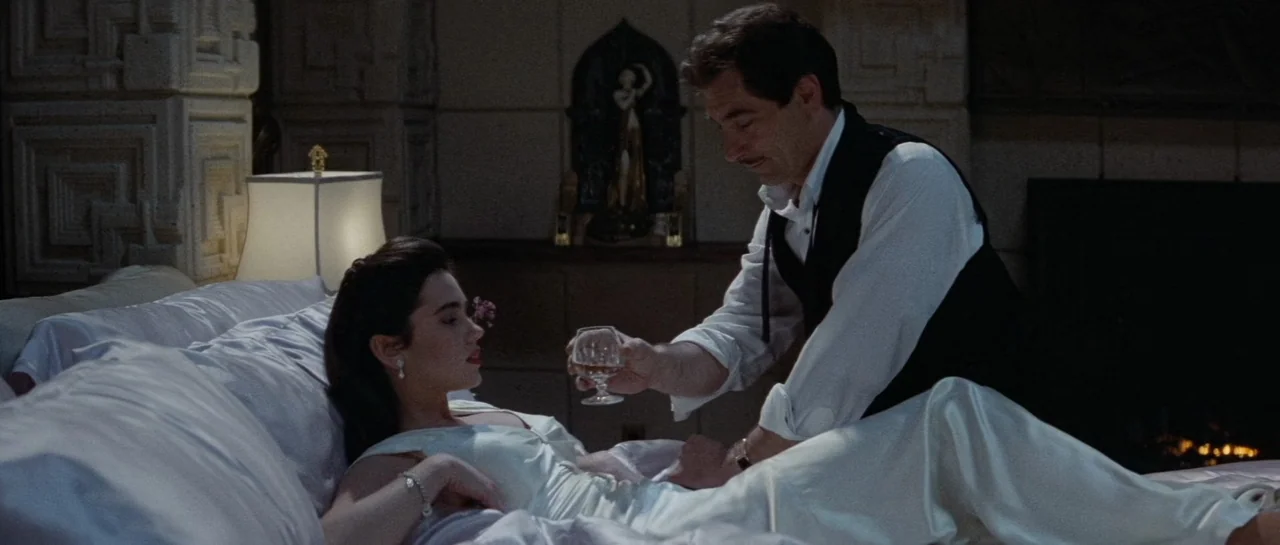 The Rocketeer (1991) movie still: medium — A dimly lit room features a bed with white silk linens and ornate wall carvings. A man leans toward a…